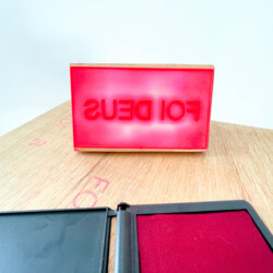 Almofada de tinta vermelha com carimbo escrito "Foi Deus" sob uma mesa de madeira