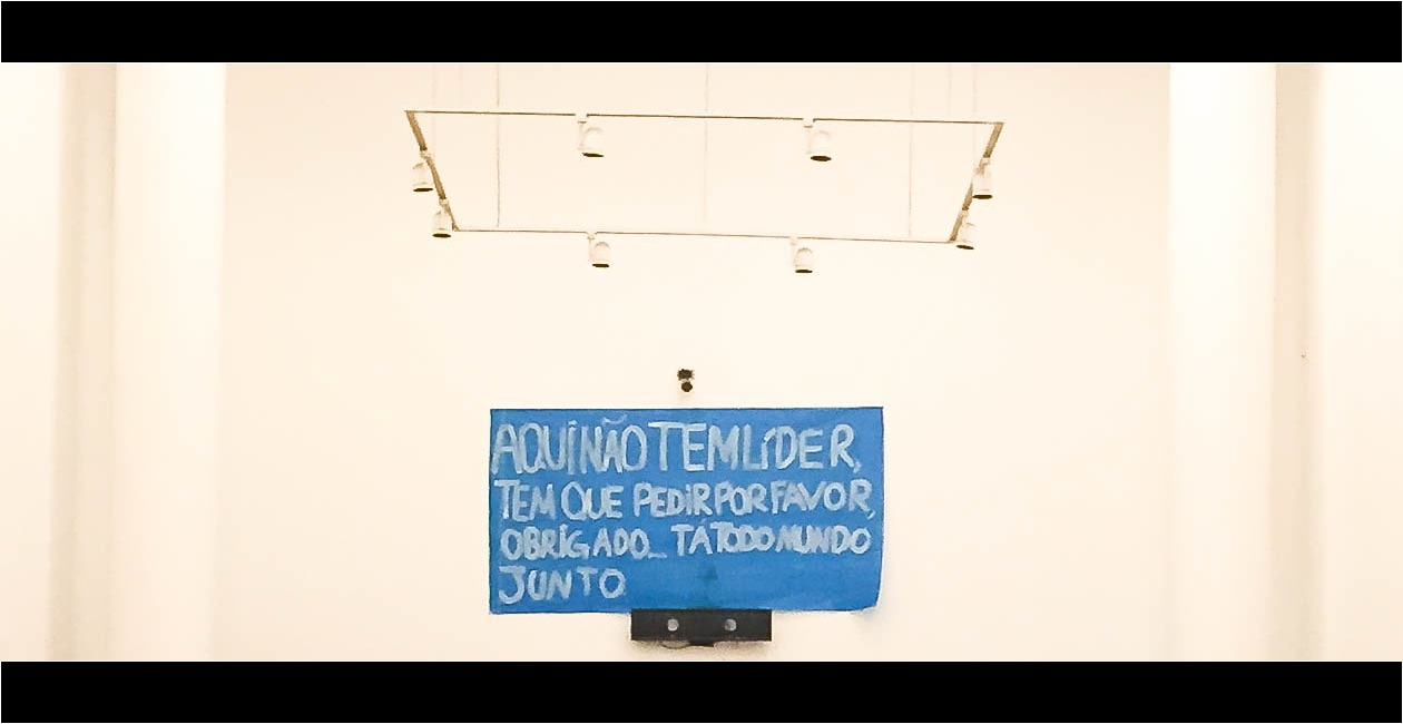 Foto de parede com papéis colados com frases, sendo uma delas "Aqui não tem líder, tem que pedir por favor, obrigado... Tá todo mundo junto."