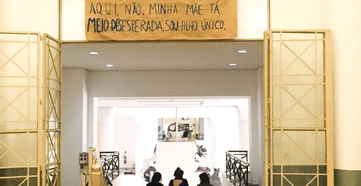 Porta aberta com salão a frente e sob ela há um papel com a frase "Não sei se posso dormir aqui, não. Minha mãe tá meio desesperada, sou filho único."